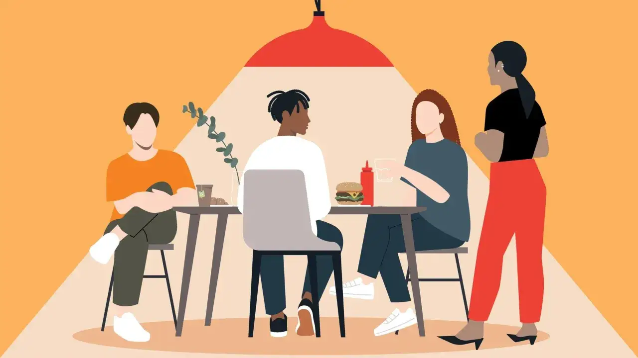 Vier Freunde treffen sich in einem Café. Zwei sitzen an einem Tisch, ein extrovertierter Mann und eine Frau. Eine Frau steht daneben, während ein Mann auf der anderen Seite sitzt.
