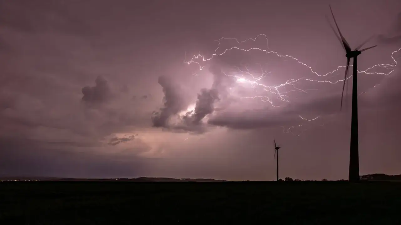 Gewaltiger Blitz zuckt über den Himmel, während zwei Windräder im Sturm stehen. Ein orkan-artiges Naturschauspiel.