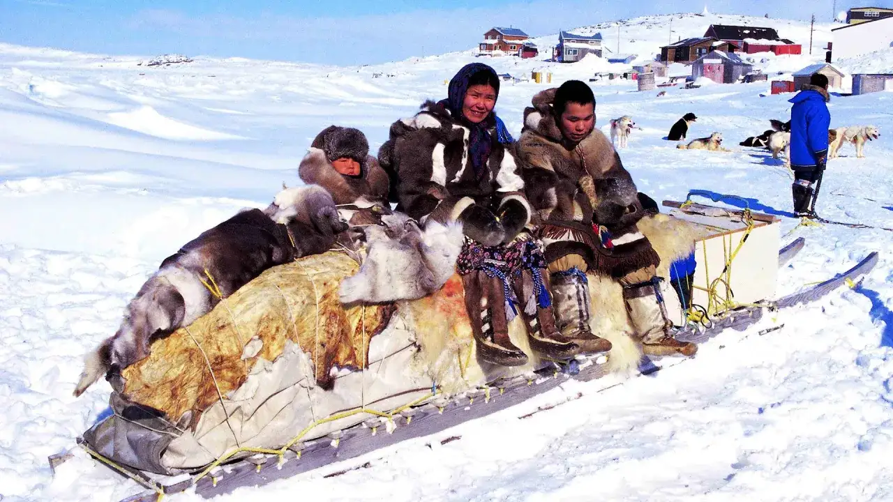 Inuit-Familie in traditioneller Kleidung auf einem Schlitten, umgeben von Hunden und schneebedeckten Häusern.