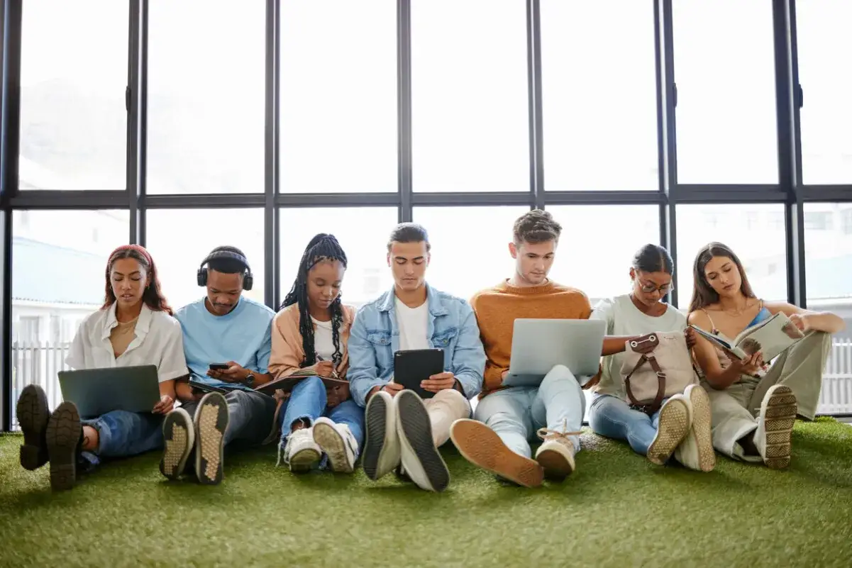 Sechs junge Leute, der gen z jahrgang, sitzen auf Kunstrasen vor einem Fenster und arbeiten an Laptops, Tablets oder lesen.