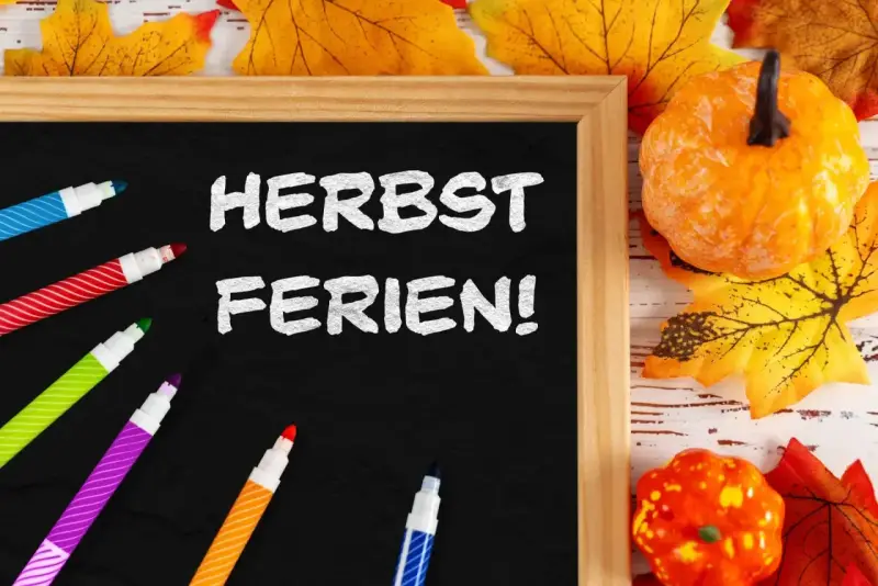 Bunte Stifte und Herbstdeko umrahmen eine Tafel mit der Aufschrift "Herbstferien!". Zeit für die Herbstferien Baden-Württemberg 2025!